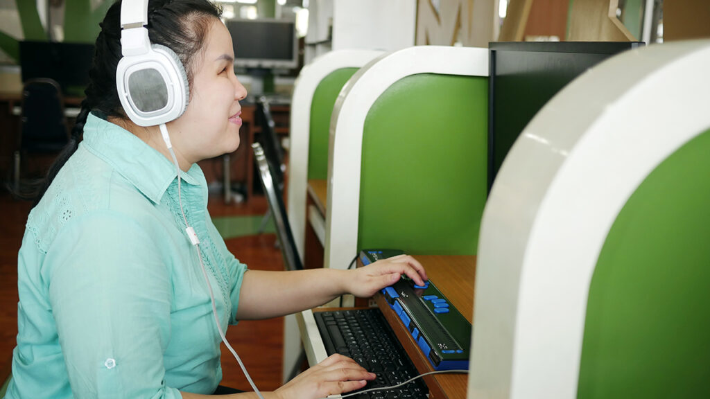 Visually-impaired woman sitting at a computer for the blind. She has white headphones on.