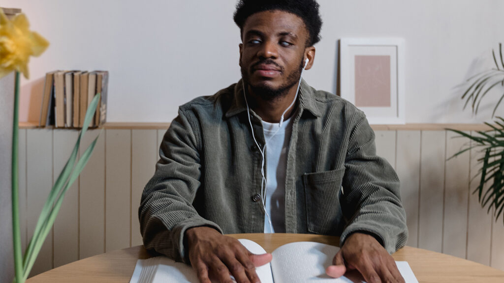 Young visually-impaired Black man wearing headphones and reading a braille book.