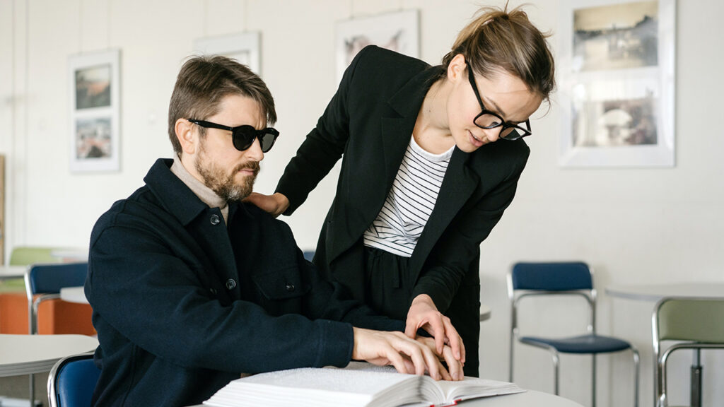 White woman training white visually-impaired white man to read braille.