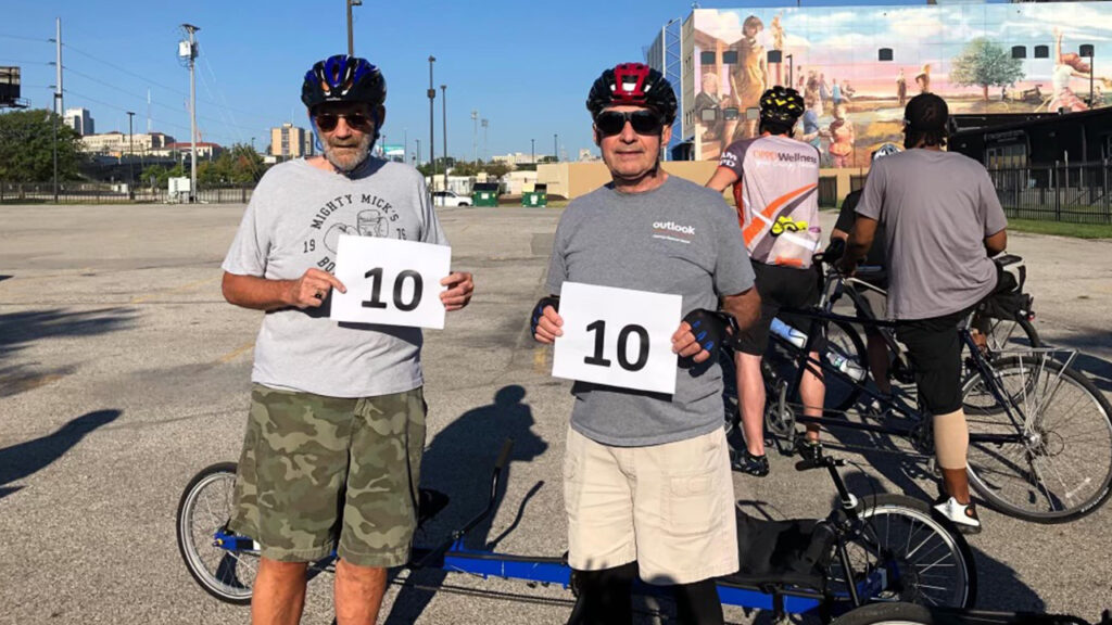 Two men in bike helmets holding up signs with the number 10 as they're about to participate in Enrichment’s adaptive sports program.