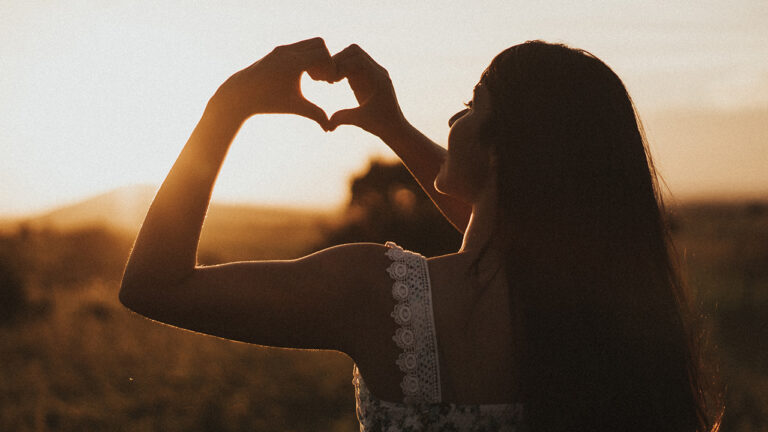 Woman with long hair with her back to the camera, holding her hands up as a heart.