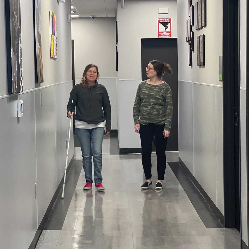 Young, white visually impaired woman walking down a hallway with a cane and another young white woman walking on her right side.