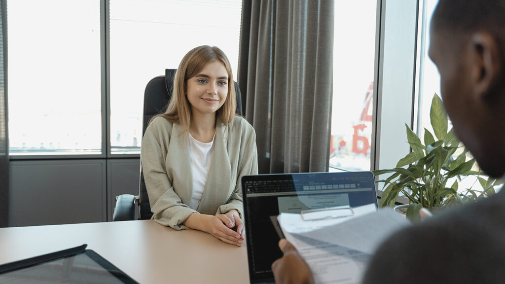 Smiling white woman sitting at a desk getting interviewed by a Black man who is reviewing her resume.