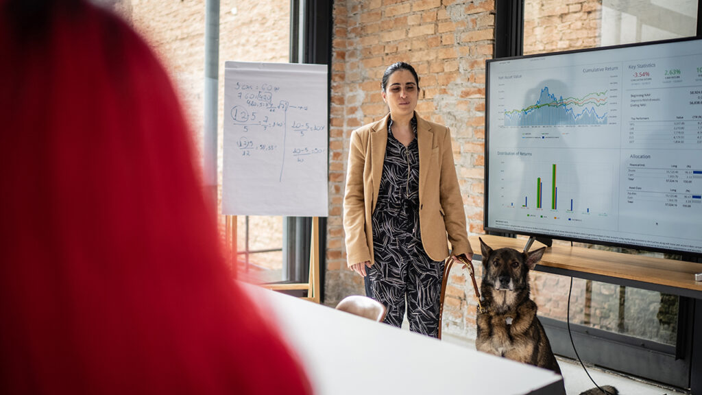 Visually-impaired woman standing in front of a TV screen with graphs on it with her German Shephard guide dog.