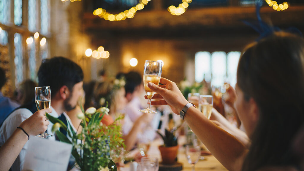 Woman out of focus and raising a toast with a glass of champagne. They're sitting at a table with other people.