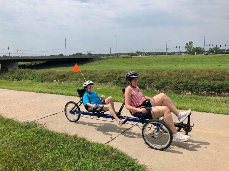 A woman and a teenage girl on a sit down recumbent tandem bike. They are smiling and pedaling on a trail. There is green grass on either side of the bike trail.