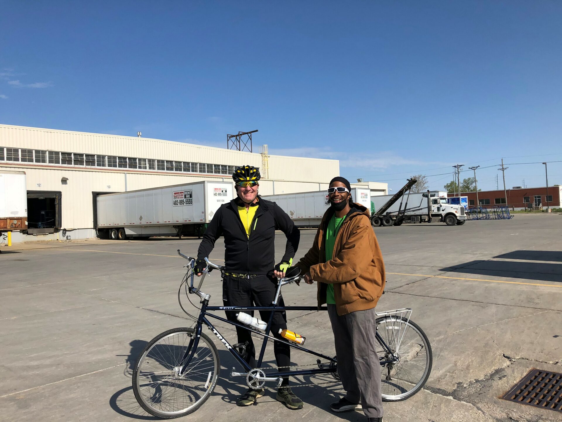 Two men standing behind a standard stand-up recumbent tandem bike. They are wearing helmets and outside in a parking lot about to take off for a ride.