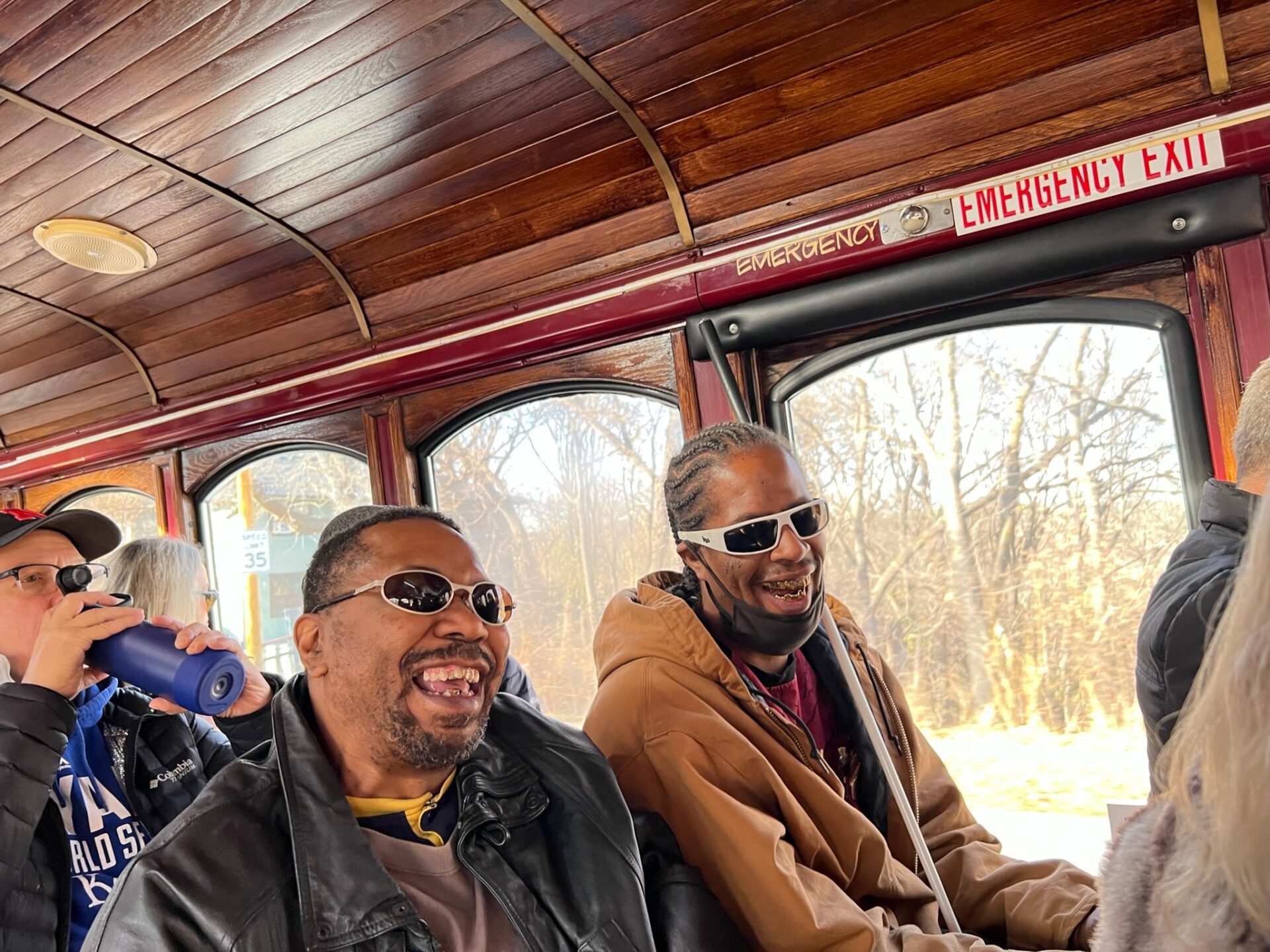 Two black men with sunglasses on sitting side by side on a bench inside Ollie the Trolley. The trolley windows and ceiling are showing in the background.