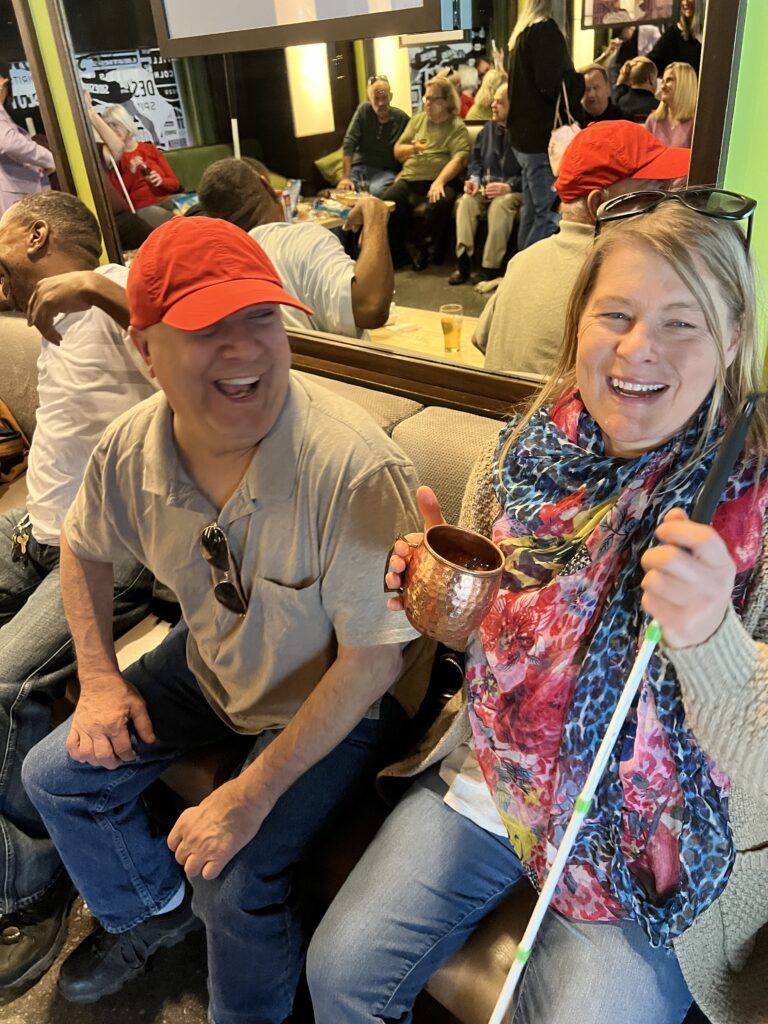 A man wearing a red hat and a lady laughing, holding a white cane are sitting on a couch in a lounge.