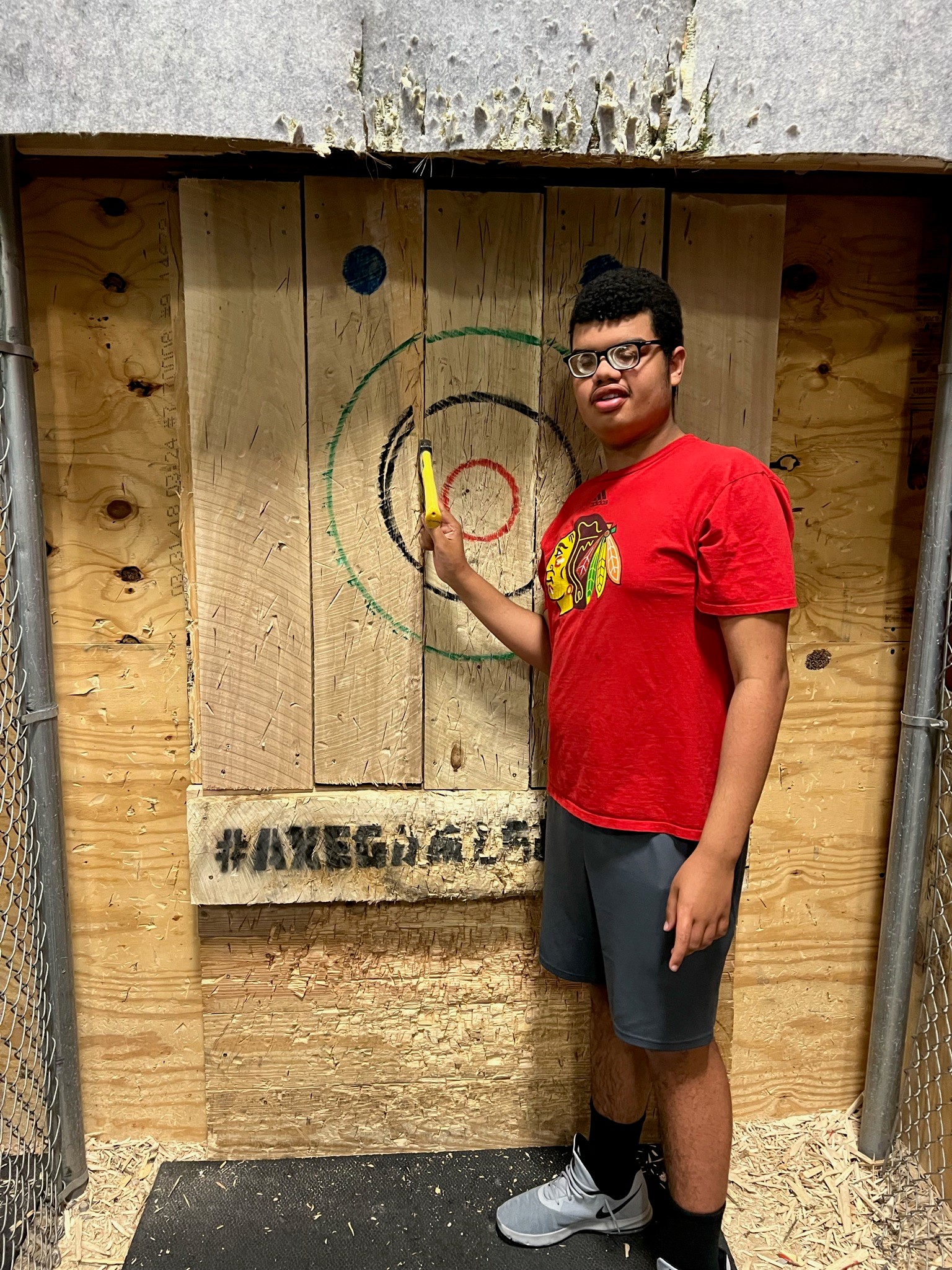 A young man wearing glasses is standing in fron of a axe throwing target. He is pointing at the axe he threw just outside of the bullseye.
