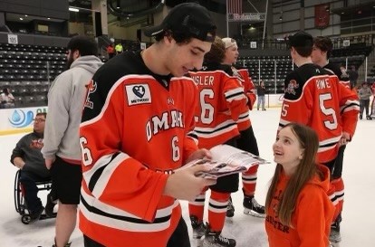 A Lancer's hockey player and a young girl are on an ice rink. The Lancer's player is wearing his orange, black and white hockey jersey and the girl is looking up at him smiling.