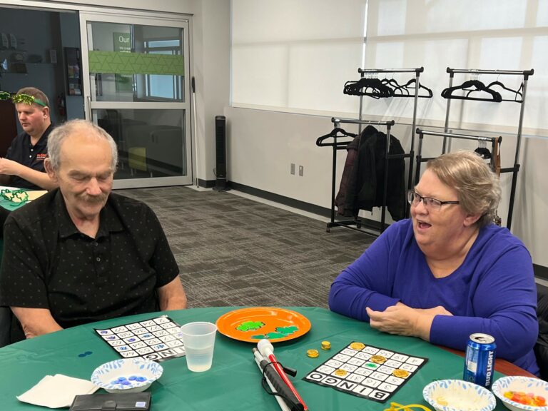 A man and a woman sitting at a table with large print bingo cards. The woman's mouth is open and she is shouting out BINGO!