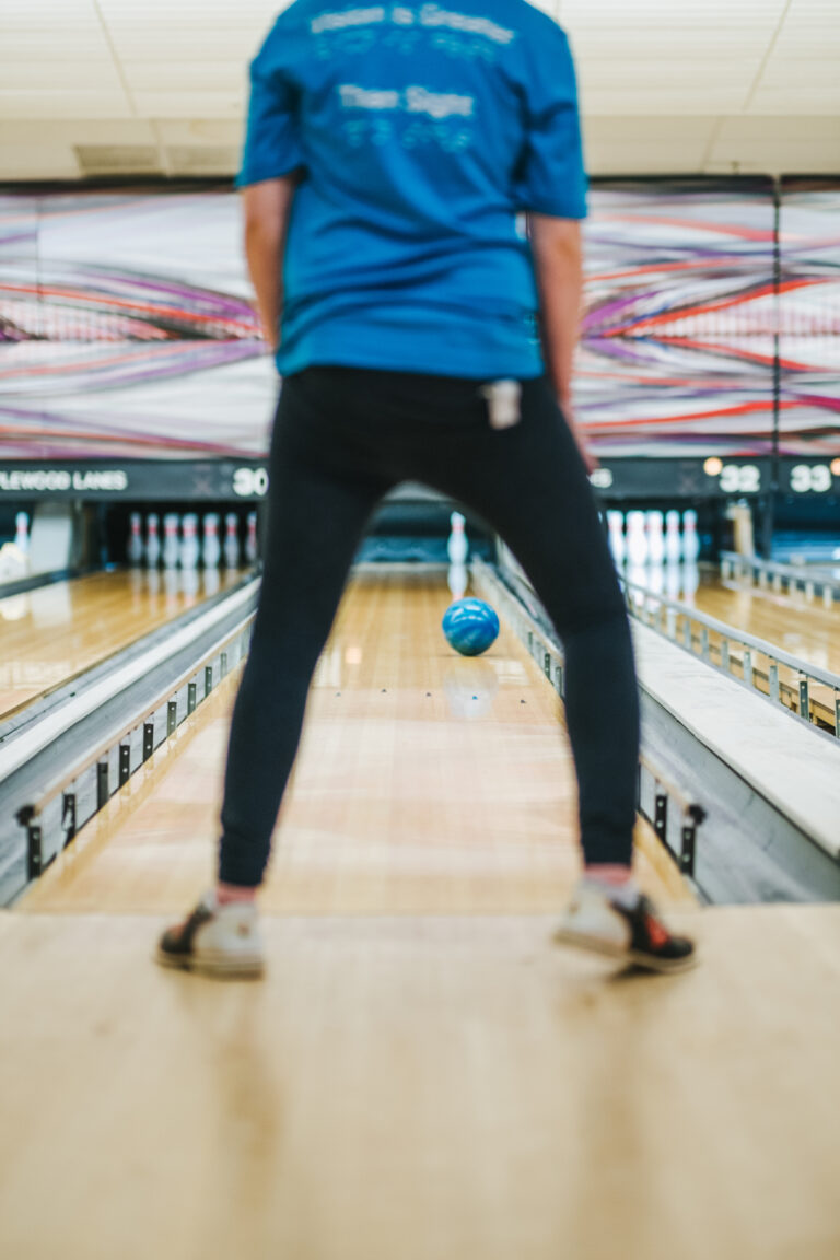 A person pictured from the back is wearing an Outlook shirt. They are faceing bowling lanes and a bowling ball is rolling down the lane they are in front of.