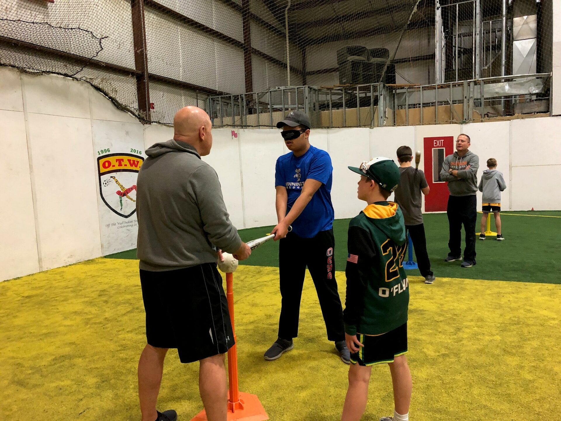 Adaptive Sports Specialist, Mike Messerole, gives a visually impaired teen some pointers on batting. He is holding a bat in his hands, lined up with the tee and ball. A sighted little league player watches them practicing.