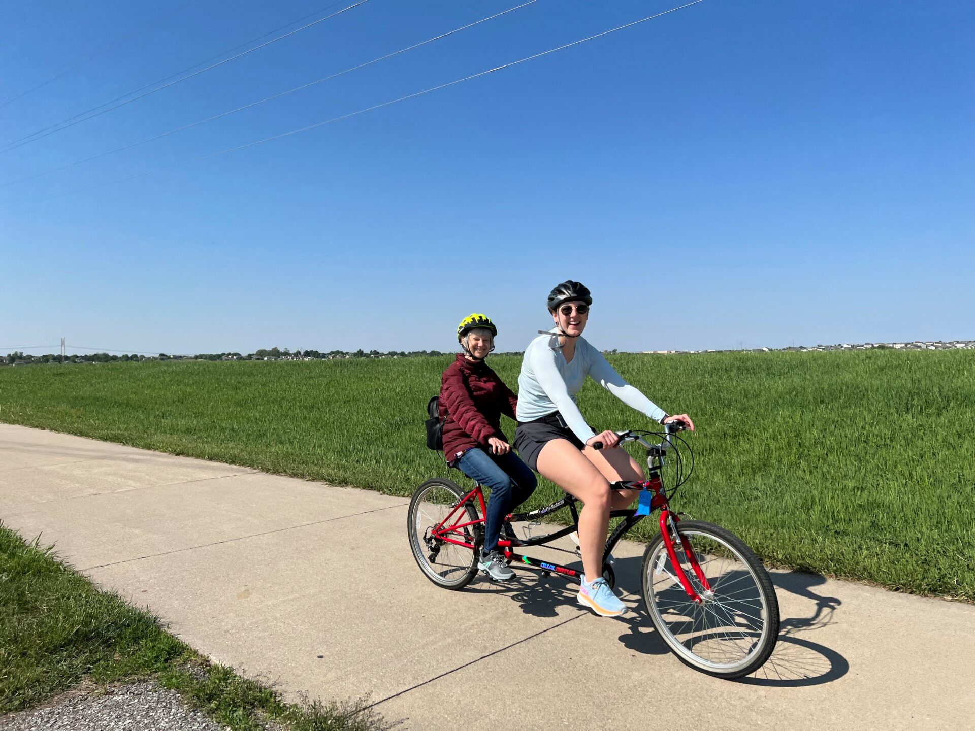 Two ladies riding a tandem bike with two seats. They are wearing helmets and smiling on a trail with grass in the background.