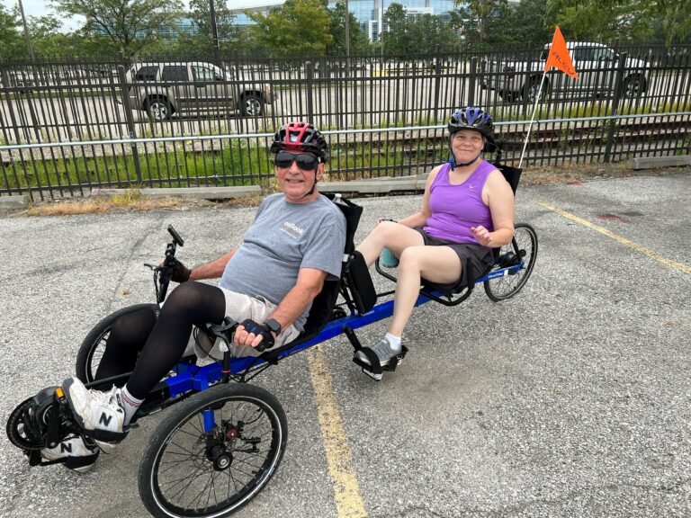 A man in the front seat of a recumbent bike with a woman in the back seat. They have helmets on and they are in a parking lot getting ready to take off for the Corporate Cycling Challenge.