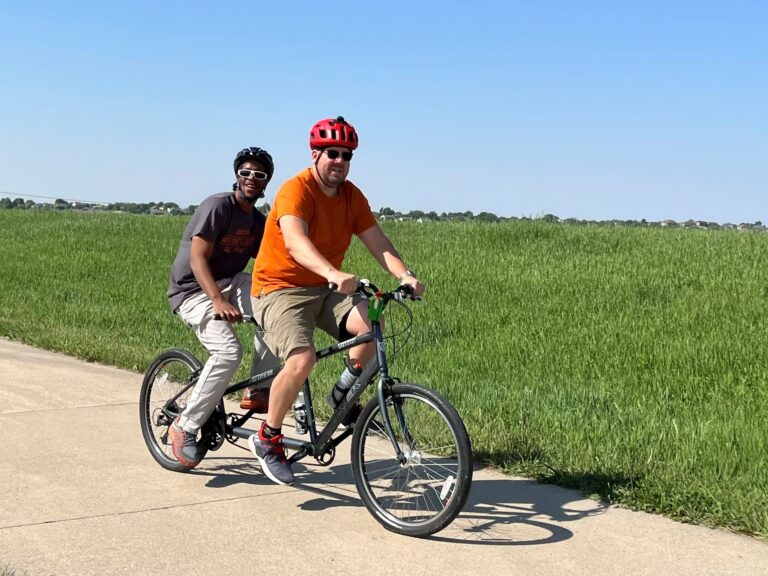 Shawn, a volunteer bike captain is on the front seat of a standard tandem bike and Mortel is on the back seat. They are in action pedaling on a path on the Keystone Trail with a grassy field in the background.