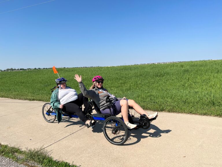 Two women wearing helmets and pedaling on a recumbent bike on a bike trail with grassy fields in the background.