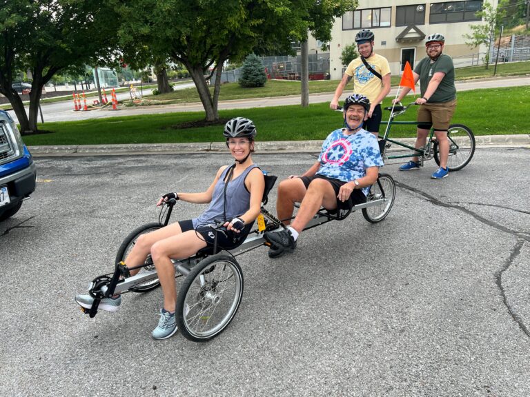 A two seated bike with a lady in the front seat and one of our regular bikers, Tim, on the back seat wearing a tie-dye shirt and a bike helmet.
