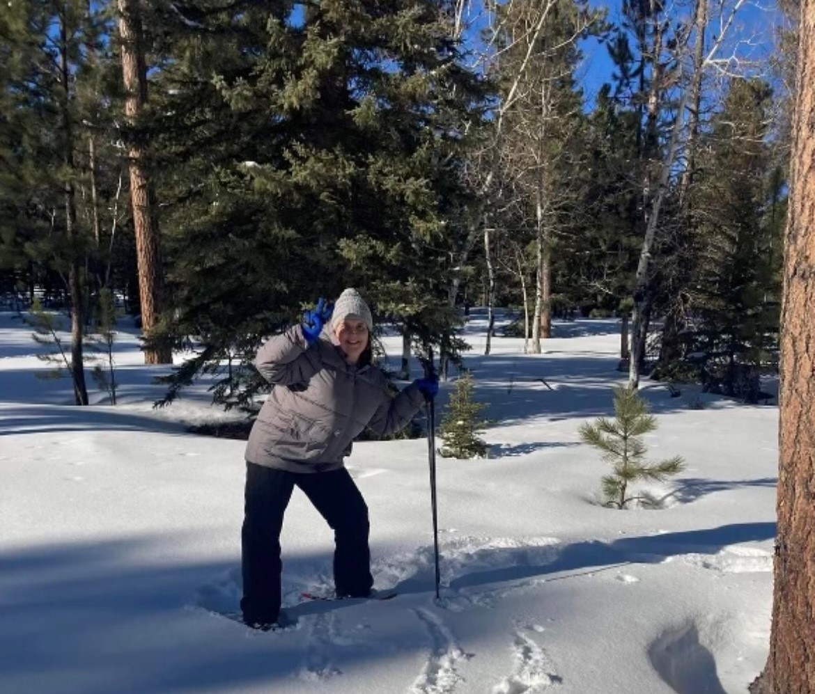 A lady wearing a hooded sweatshirt and coat. She has on snowshoes and is holding ski poles as she hikes across a snowy field with trees in the background.
