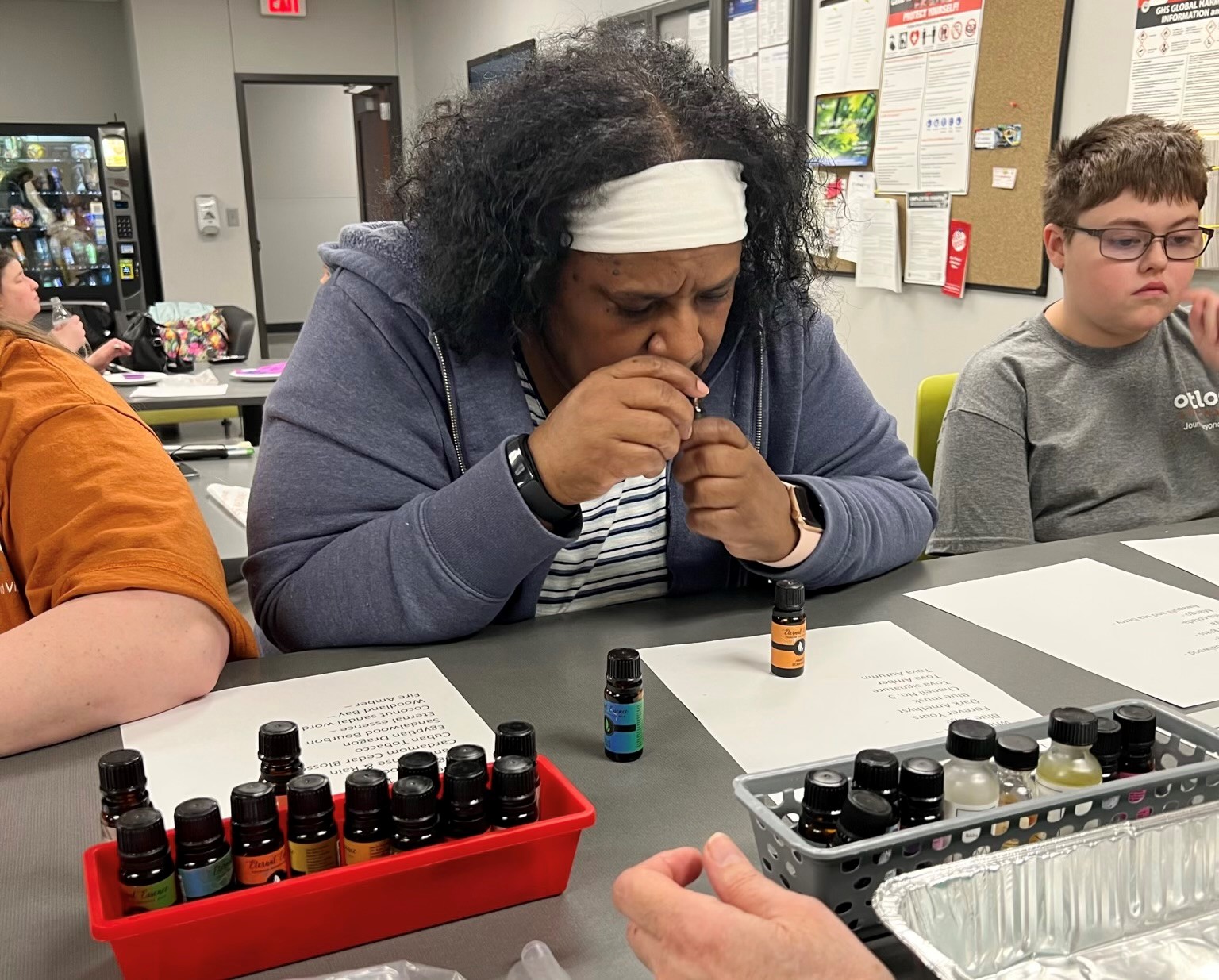 A black woman standing at a table filled with scented oils. Her head is bent down and her nose is near the scented oil bottle she is holding up.