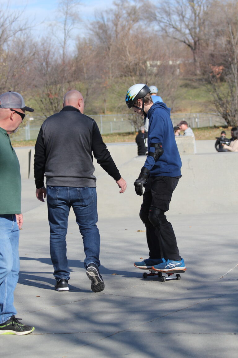 A teenager wearing a helmet, on a skateboard at an outdoor skate park. An adaptive sports specialist is next to him as he balances on the skateboard.