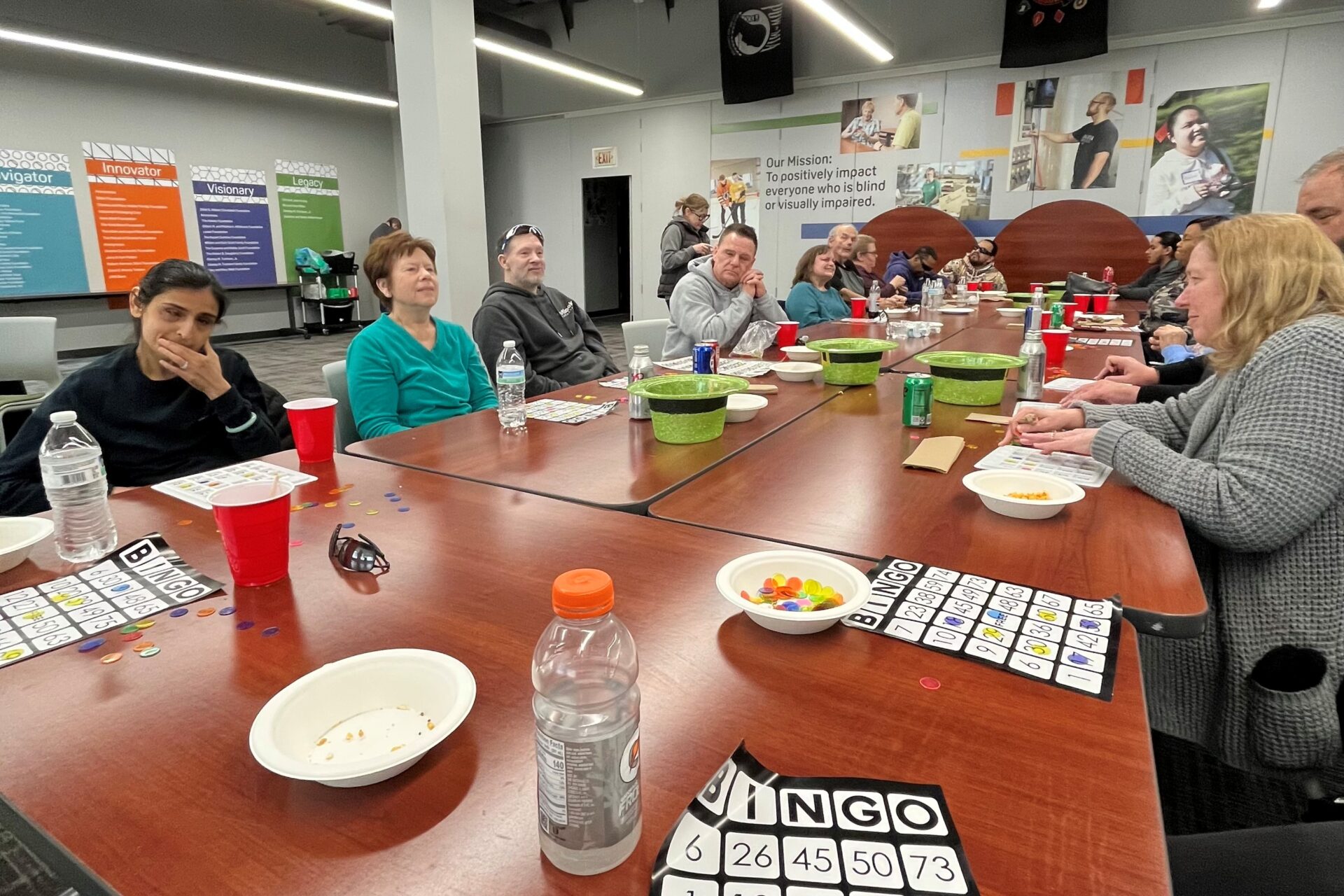 a very long rectangle table with people all around it. There are refreshments and bingo cards on the table. Kristal is using her fingers to read a braille bingo card.